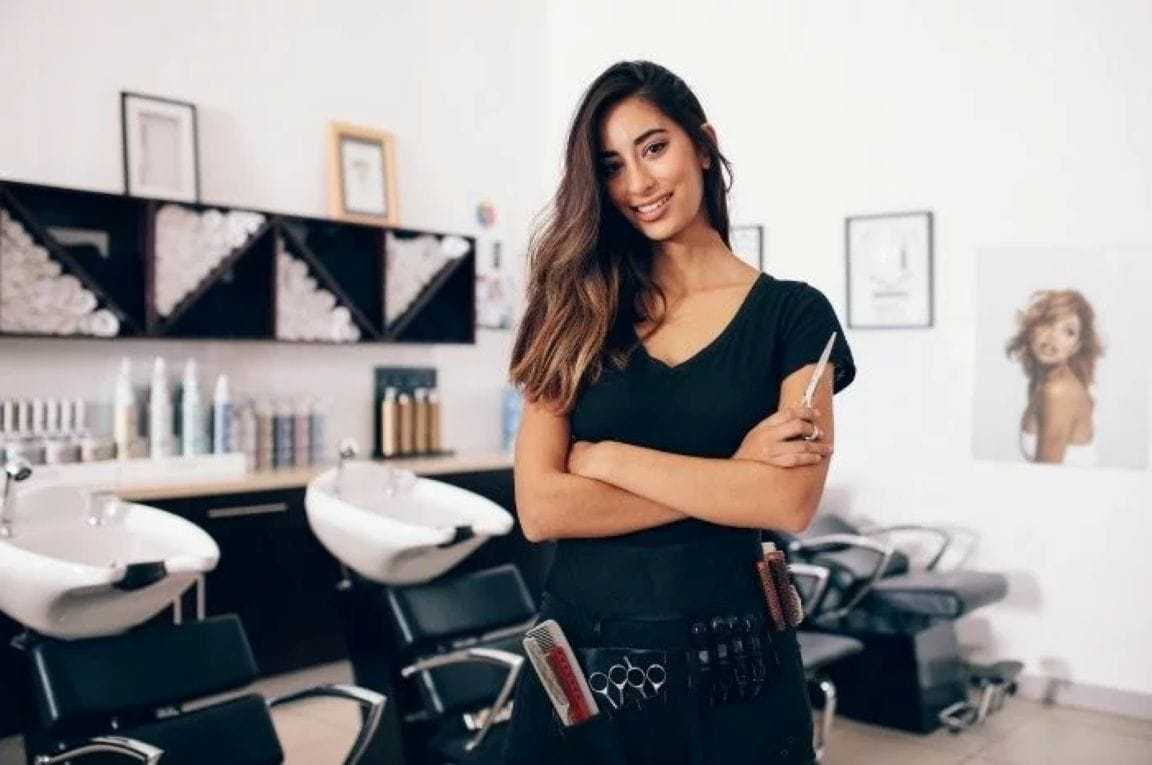 Hairstylist standing in a salon, smiling, with styling tools in her apron.